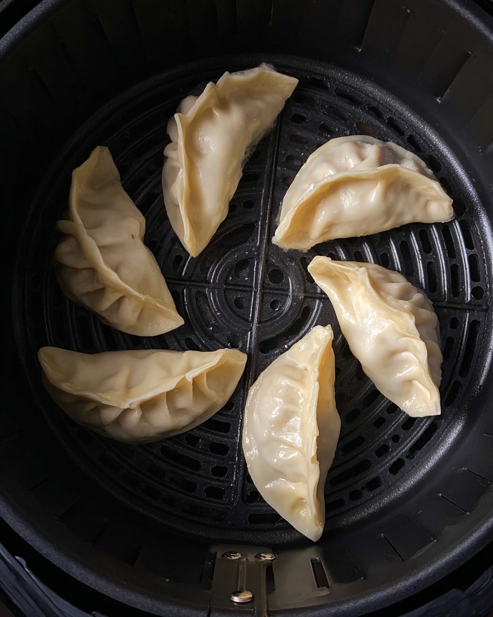 Six uncooked dumplings with a pale, smooth dough surface are arranged in a circle inside a black air fryer basket. Each dumpling is half-moon shaped with crimped edges creating folds, showing slightly different folds on each piece. The dumplings have a slight shine from moisture or oil on their surface, resting on the dark textured basket with its circular and line patterns visible under them. Photo taken with an iphone --ar 4:5 --v 7