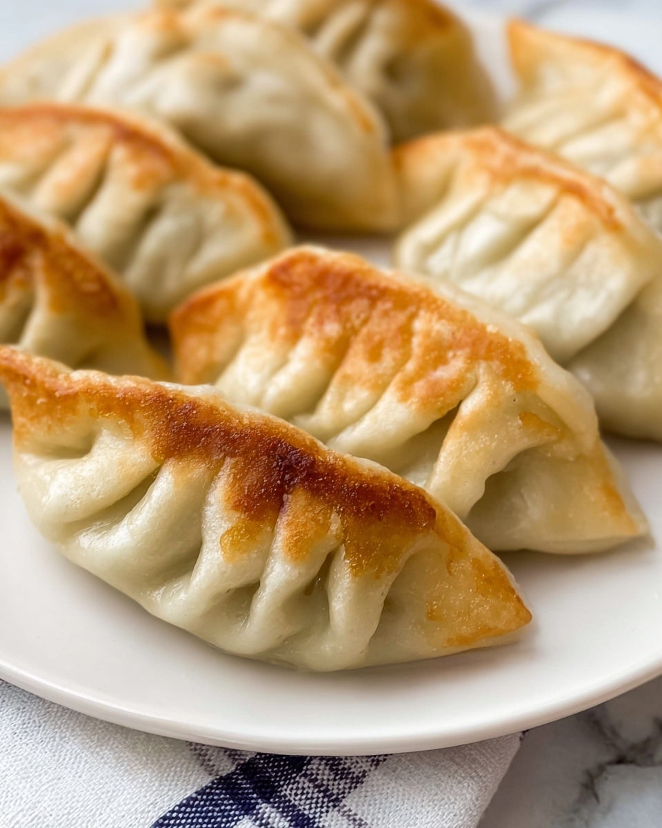 The image shows five fried dumplings arranged closely together on a white plate. Each dumpling has a golden brown edge with a soft, slightly shiny surface that is light beige in color. The dumplings have distinct pleated folds on their tops, giving them a textured, crinkled look. They are resting on a white marbled surface with a white and blue checkered cloth partially visible beneath the plate. photo taken with an iphone --ar 4:5 --v 7
