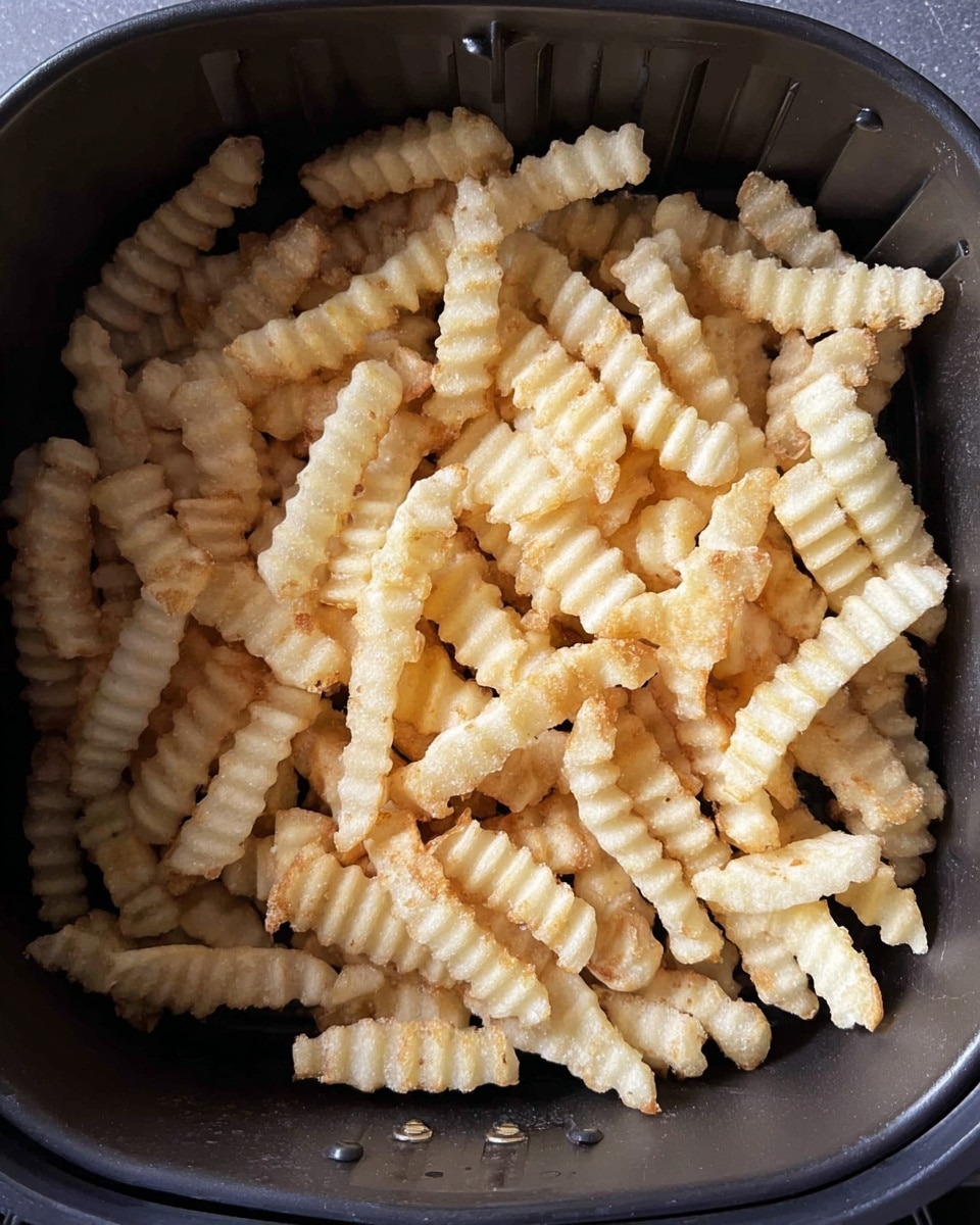 A black air fryer basket filled with a single layer of uncooked crinkle-cut fries. The fries are light beige in color, with a frosty, frozen texture and ridged edges. They are evenly spread, touching each other but not piled up, showing the dark interior of the air fryer beneath. The edges of the fries show slight variation in size and shape, adding texture to the overall image. photo taken with an iphone --ar 4:5 --v 7