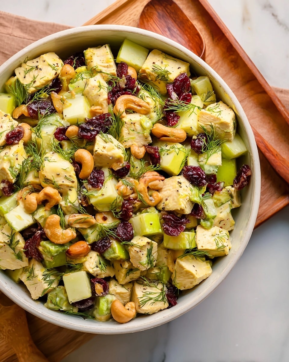 A bowl filled with a mixed salad showing several layers: the bottom layer has large chunks of light yellow cooked chicken pieces sprinkled with small green dill leaves; mixed in are medium-sized pale green apple pieces and light green celery slices. Scattered throughout are golden brown cashew nuts and dark reddish-brown dried cranberries, along with small green onion slices. The bowl is white and round, placed on a white marbled textured surface partially covered by a wooden board. Photo taken with an iphone --ar 4:5 --v 7