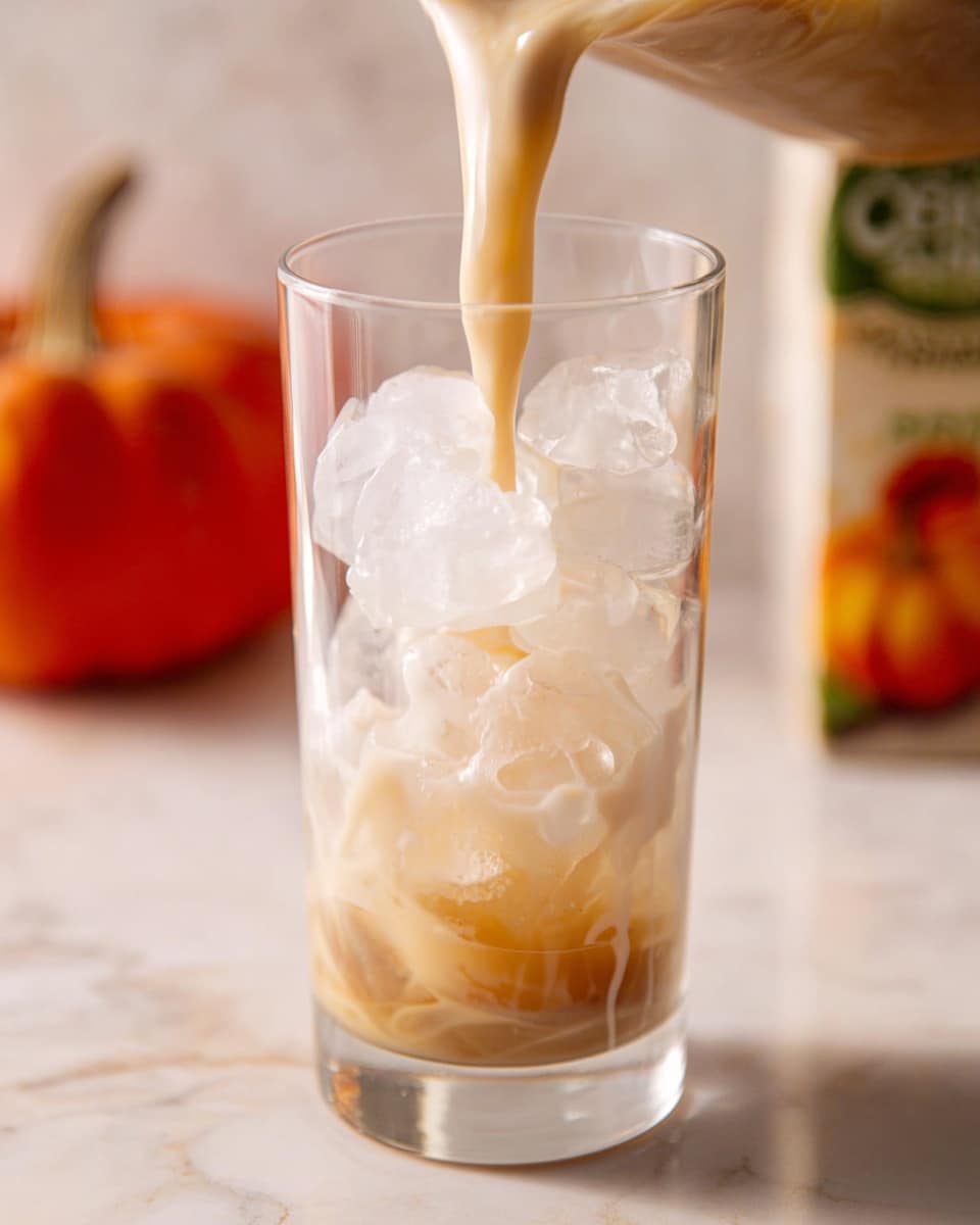 A clear tall glass filled with large, clear ice cubes forms the bottom layer, showing a cold, refreshing base. Light beige liquid is being poured from above, creating a smooth stream that hits the ice and splashes gently, starting to fill the glass. The background features a white marbled texture with a blurred carton showing a pumpkin image to the right. The overall scene suggests a creamy cold drink being prepared. photo taken with an iphone --ar 4:5 --v 7