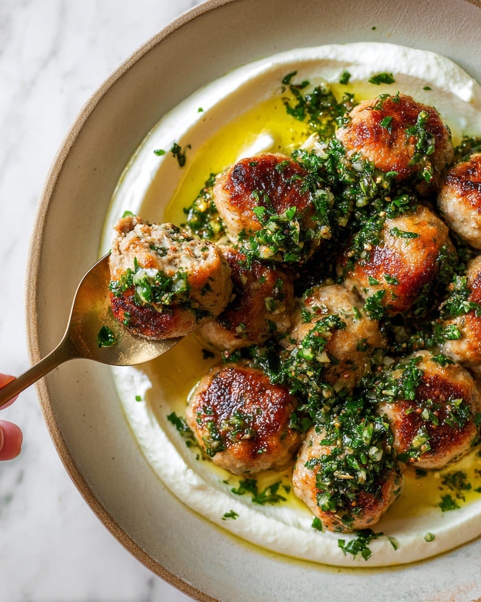 The image shows a close-up of a white plate with about ten golden brown meatballs arranged in a cluster. Each meatball has a slight crispy surface and is topped with bright green chopped herbs mixed with tiny bits of garlic or onion. Below the meatballs, there is a thick, creamy white sauce spread in a loose, uneven circle around the plate’s center, with some yellowish oil pooling near the center. A bronze spoon holds one meatball, scooping it slightly from the group, with the woman's hand barely visible holding the spoon. The background is a white marbled texture. Photo taken with an iphone --ar 4:5 --v 7