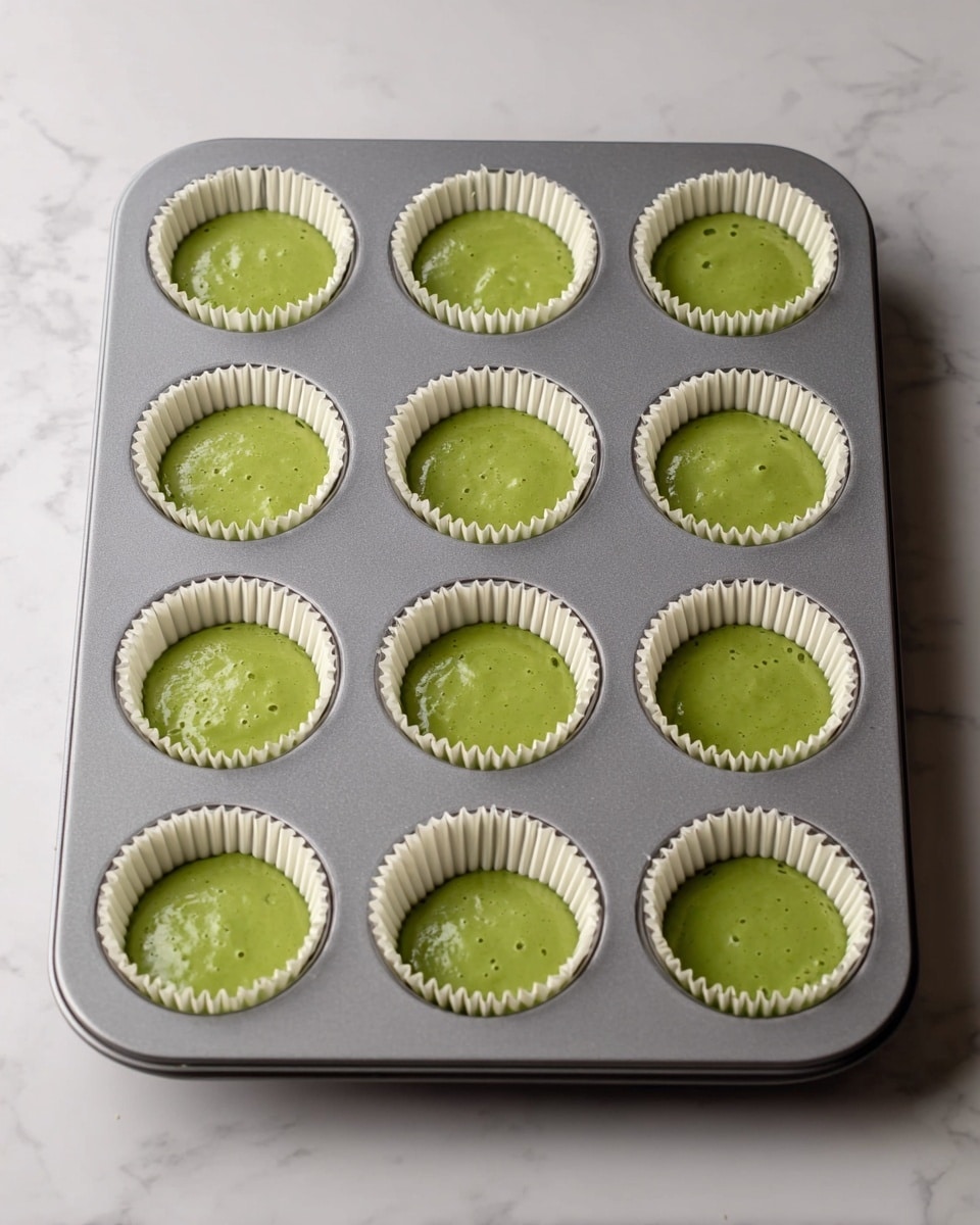 A gray muffin tray with twelve white paper liners is placed on a white marbled surface. Each liner is filled with a smooth green batter, evenly spread and filling about half the depth of each cup. The texture of the batter looks creamy and thick, with subtle bubbles on the surface indicating it is ready to bake. Photo taken with an iphone --ar 4:5 --v 7