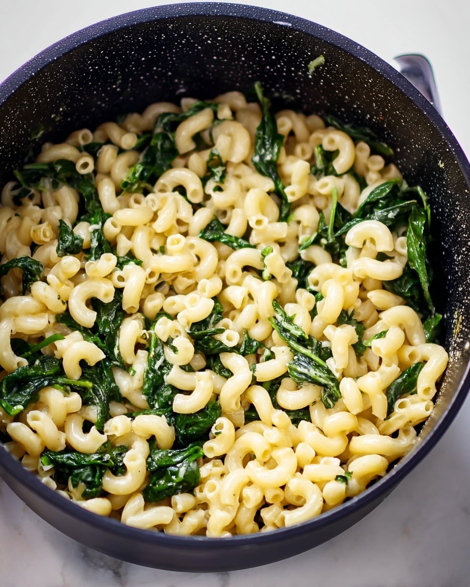 The image shows a black cooking pot filled with cooked elbow macaroni pasta and leafy green spinach mixed in evenly. The pasta is pale yellow and smooth, while the spinach is dark green and wilted, creating a nice contrast. Small water droplets are visible on the pot's sides, giving a fresh, just-cooked feel. The pot sits on a white marbled surface photo taken with an iphone --ar 4:5 --v 7