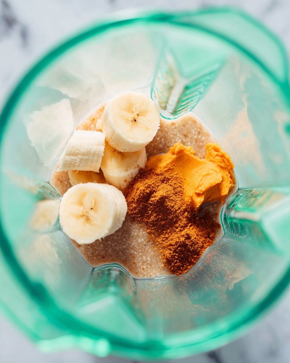 Inside a clear blender, there are four visible layers of ingredients from bottom to top: the base is a grainy light brown powder, followed by a bright orange thick paste, then a smaller dusting of a darker brown powder scattered unevenly, and on the very top, five slices of banana with pale yellow color and soft texture arranged loosely. The blender walls are transparent, showing a slight greenish tint from lighting. The background is a white marbled texture. photo taken with an iphone --ar 4:5 --v 7