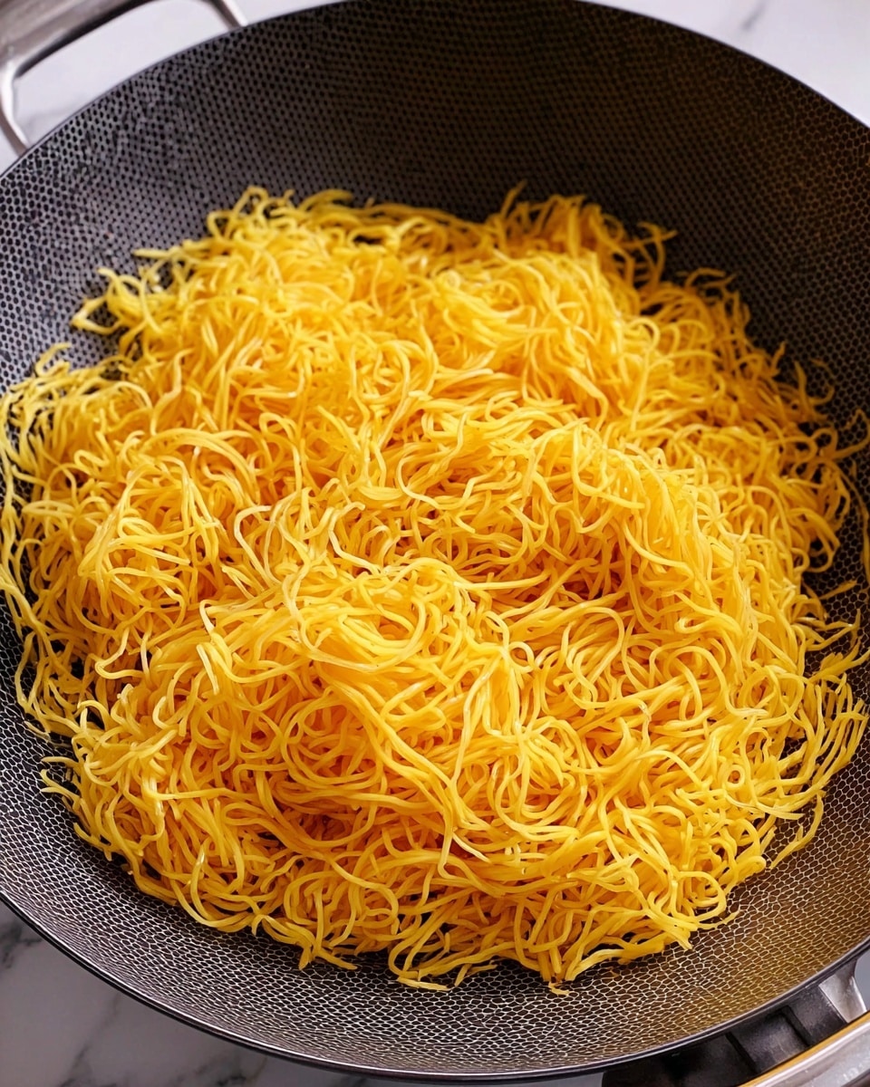 A wok filled with a large layer of cooked yellow noodles that look soft and slightly shiny, with thin strands tangled together closely covering the whole bottom of the wok, which has a dark gray hexagonal texture on the inside, placed on a white marbled surface photo taken with an iphone --ar 4:5 --v 7