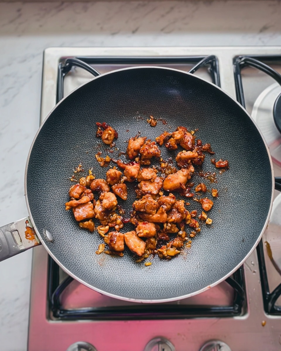 A close-up of a dark gray frying pan with a textured inside surface, sitting on a stove with silver metal parts and a light pink ring around the burner. Inside the pan are small pieces of cooked chicken with a shiny, brown glaze and bits of fried garlic or onion scattered around. The background is a white marbled texture surface. photo taken with an iphone --ar 4:5 --v 7