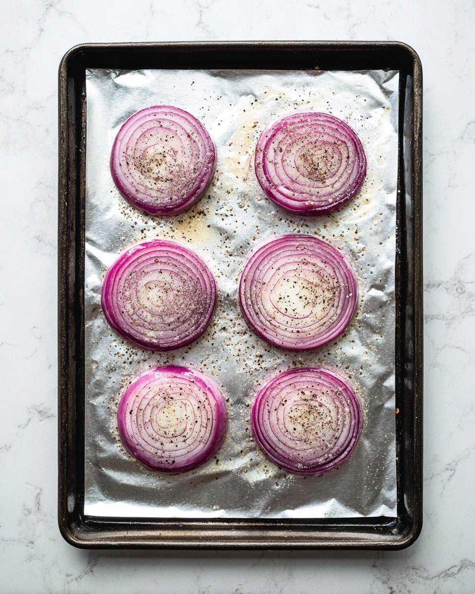 The image shows six round slices of red onion arranged in two columns and three rows on a silver foil covering a dark baking tray. Each onion slice is topped with coarse salt and cracked black pepper, with visible seasoning spread unevenly across the purple and white layers of the onions. The foil has slight crinkles and light oil stains around the onion slices, adding texture. The tray sits on a white marbled surface, creating a clean and bright background. photo taken with an iphone --ar 4:5 --v 7