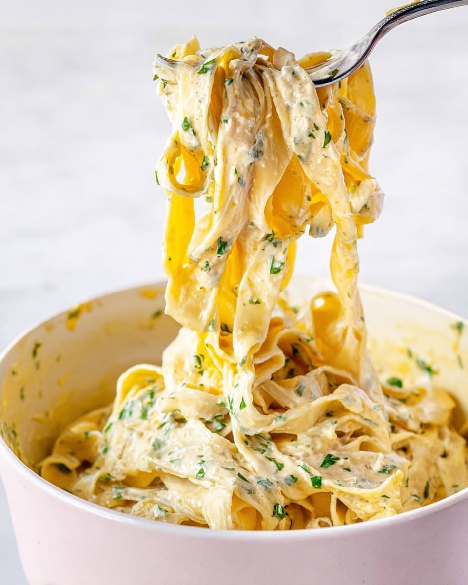 The image shows wide, flat pasta noodles covered in a creamy, pale yellow sauce with green herb bits scattered throughout. The noodles are lifted midair with a fork, showing their soft and smooth texture, while more pasta rests at the bottom of a deep white bowl. The sauce has a thick and rich look, coating every noodle evenly. The background is a white marbled surface. Photo taken with an iphone --ar 4:5 --v 7