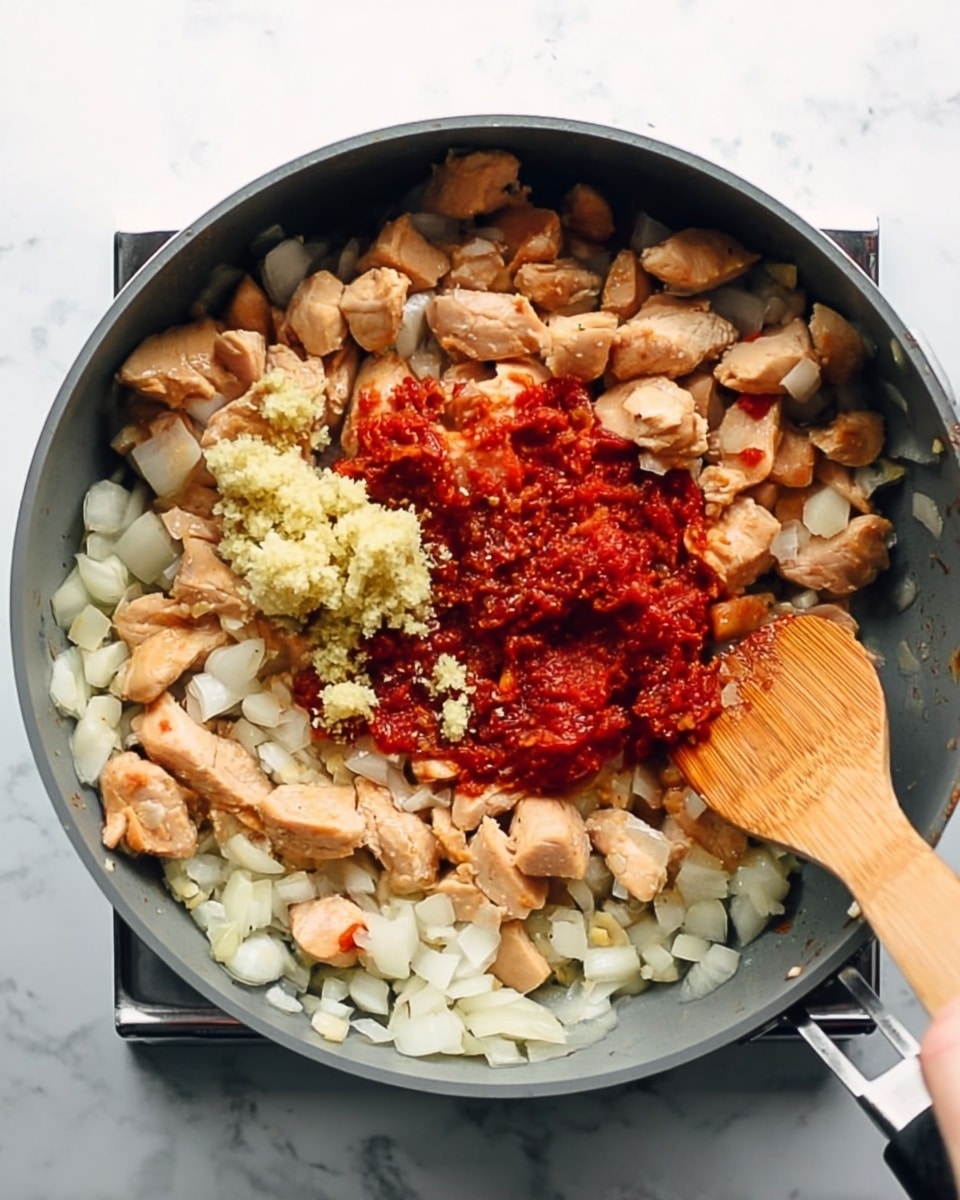 A grey pan filled with cooked light brown chicken pieces layered evenly with white diced onions, a mound of reddish tomato paste and small clumps of pale yellow minced garlic placed on top in the center; a woman's hand is holding a wooden spatula stirring the ingredients. The background surface is white with a marbled texture, photo taken with an iphone --ar 4:5 --v 7