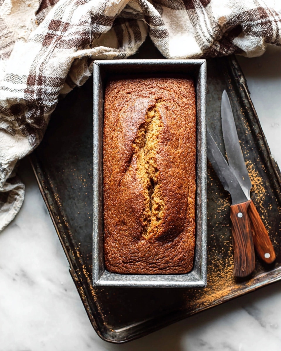 A rectangular loaf cake with a golden-brown crust sits inside a metal baking pan. The cake’s surface is slightly cracked with a textured top that shows a mix of darker and lighter brown shades, giving it a rustic, homemade look. The pan rests on a dark metal tray sprinkled with crumbs and powder, beside two wooden-handled knives placed to the right. In the upper left corner, a white and brown plaid cloth is casually folded, all set on a white marbled background. photo taken with an iphone --ar 4:5 --v 7