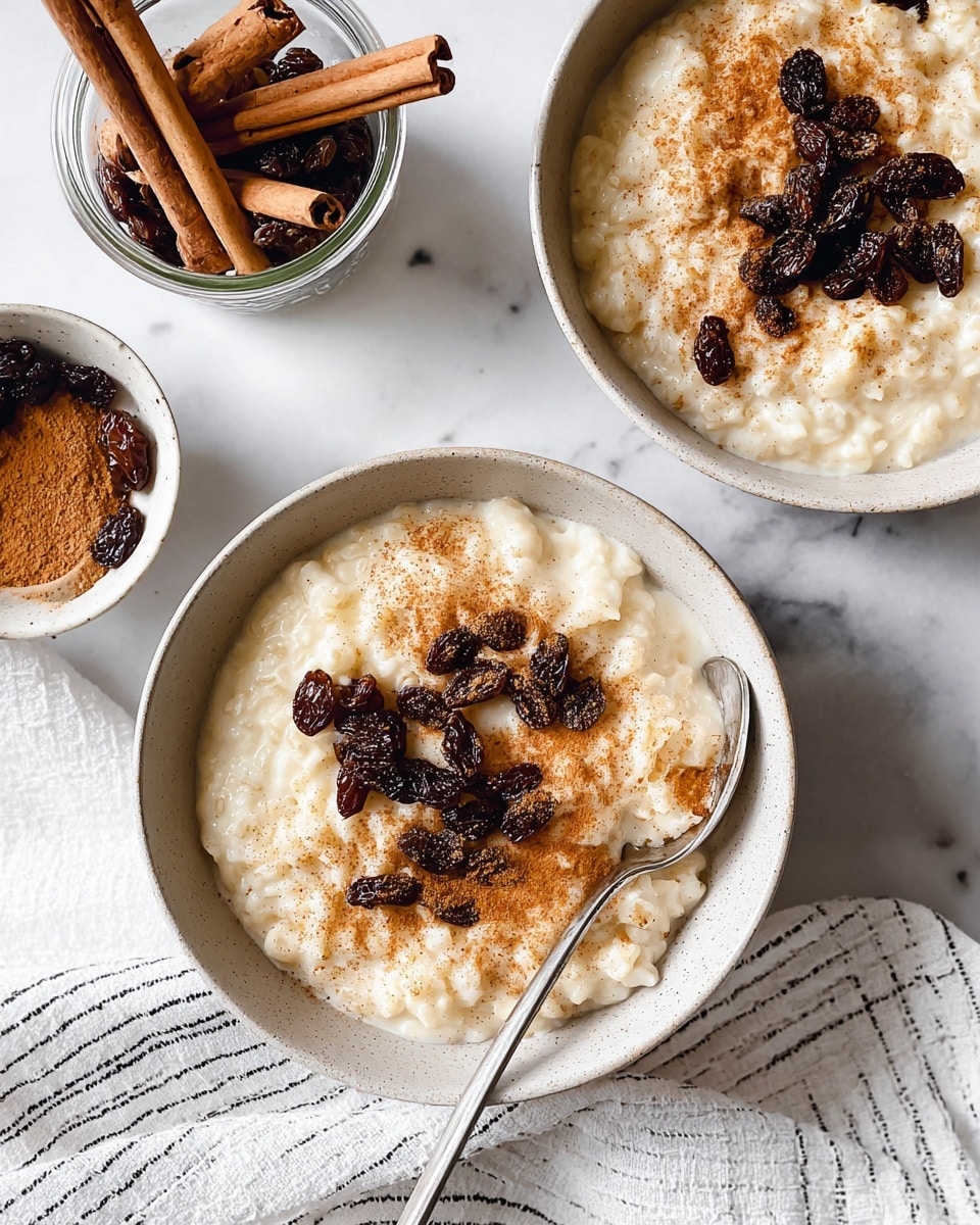 The image shows two bowls of creamy rice pudding with raisins on a white marbled surface. Each bowl has one main layer of thick, pale cream rice pudding mixed with dark brown raisins. On top, there is a sprinkle of cinnamon powder giving a warm, reddish-brown color in the center. One bowl is partially visible in the upper right corner, while the main bowl at the center has a silver spoon resting on its edge in a natural position. Next to the lower bowl, there is a white cloth with thin black stripes. Also visible are a jar with cinnamon sticks and a small bowl with more raisins, both placed on the white marbled surface. Photo taken with an iphone --ar 4:5 --v 7