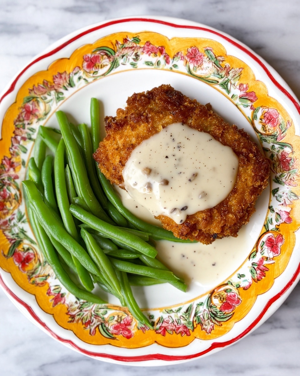 The dish shows one large crispy fried piece with a rough, golden-brown textured crust, topped with a smooth, creamy white layer of gravy that has small black pepper specks, centered on a white plate with colorful orange, yellow, red, and green floral patterns around the edges. On the top right side of the plate, there is a neat pile of glossy, bright green beans with smooth surfaces. The whole setup is placed on a white marbled surface. Photo taken with an iphone --ar 4:5 --v 7