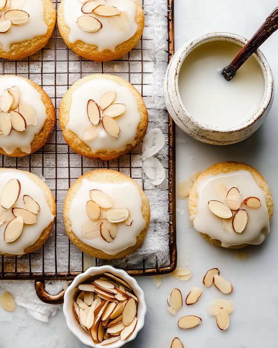 The image shows a cooling rack with eight soft round cookies placed on it, each topped with a smooth, white frosting layer that gently drips over the edges. On top of the frosting, there are three almond slices arranged like petals on each cookie, adding a light brown contrast with their thin and slightly textured look. Below and to the left of the rack, a small white bowl holds more almond slices with brown edges. Towards the back right of the rack, a rustic white bowl contains a white liquid with a dark brown spoon inside. The entire scene is set on a white marbled surface, creating a clean and bright background. photo taken with an iphone --ar 4:5 --v 7