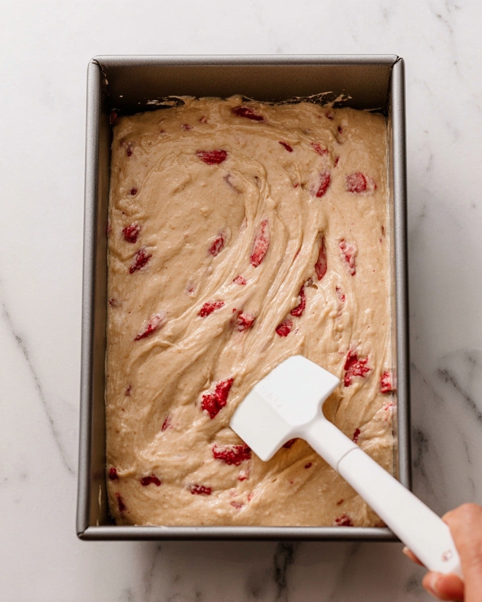 A close-up view of a metal baking pan filled with thick, light beige batter that has visible small red strawberry pieces spread throughout. A white spatula with a woman's hand holding it is partially dipped into the batter, swirling the mixture. The pan is placed on a white marbled surface. photo taken with an iphone --ar 4:5 --v 7