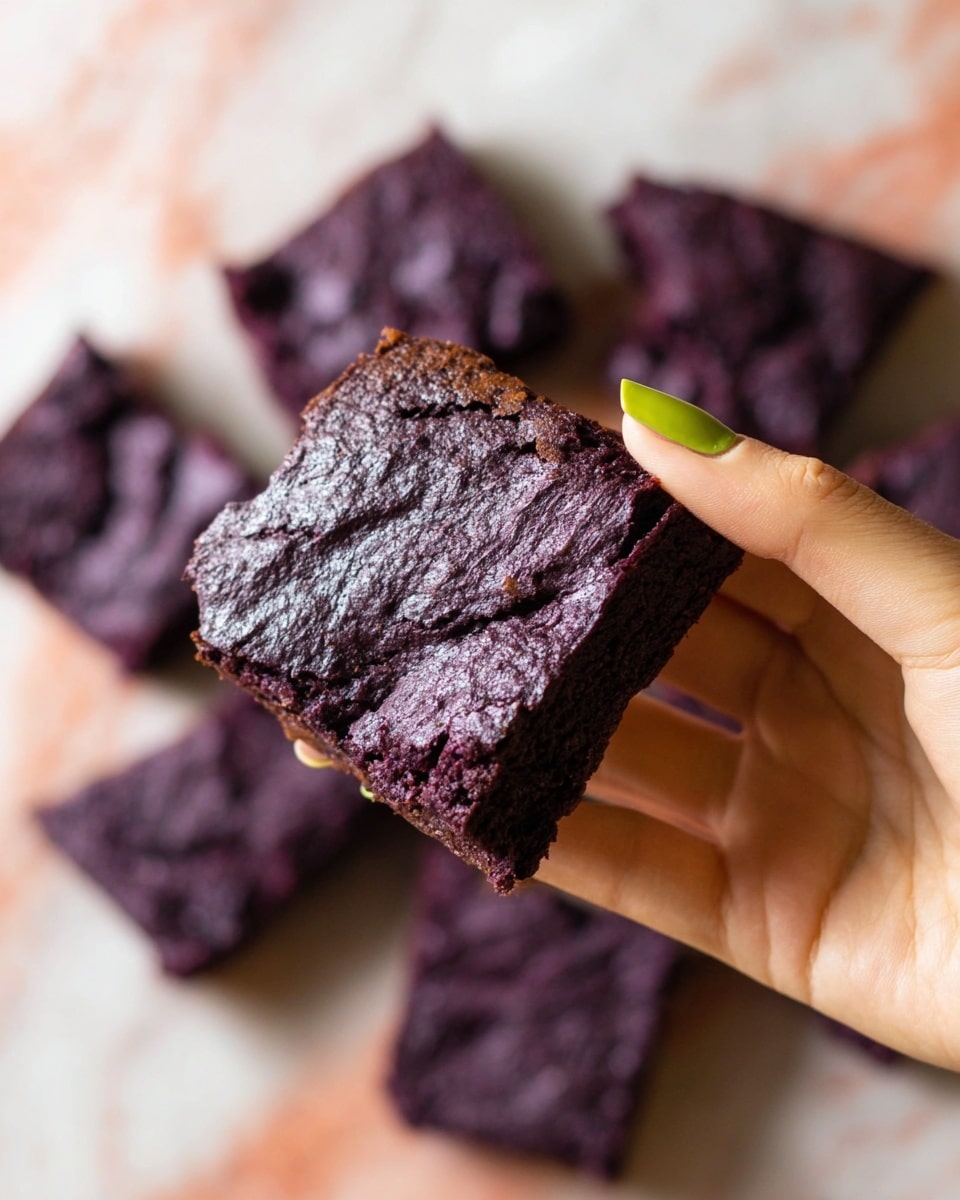 A woman's hand with green nail polish is holding a square piece of dark purple brownie with a cracked, slightly shiny top layer, showing a dense and moist interior. In the background, several more pieces of the same purple brownies are spread out on a white marbled surface, softly blurred to keep focus on the piece in the woman's hand. photo taken with an iphone --ar 4:5 --v 7