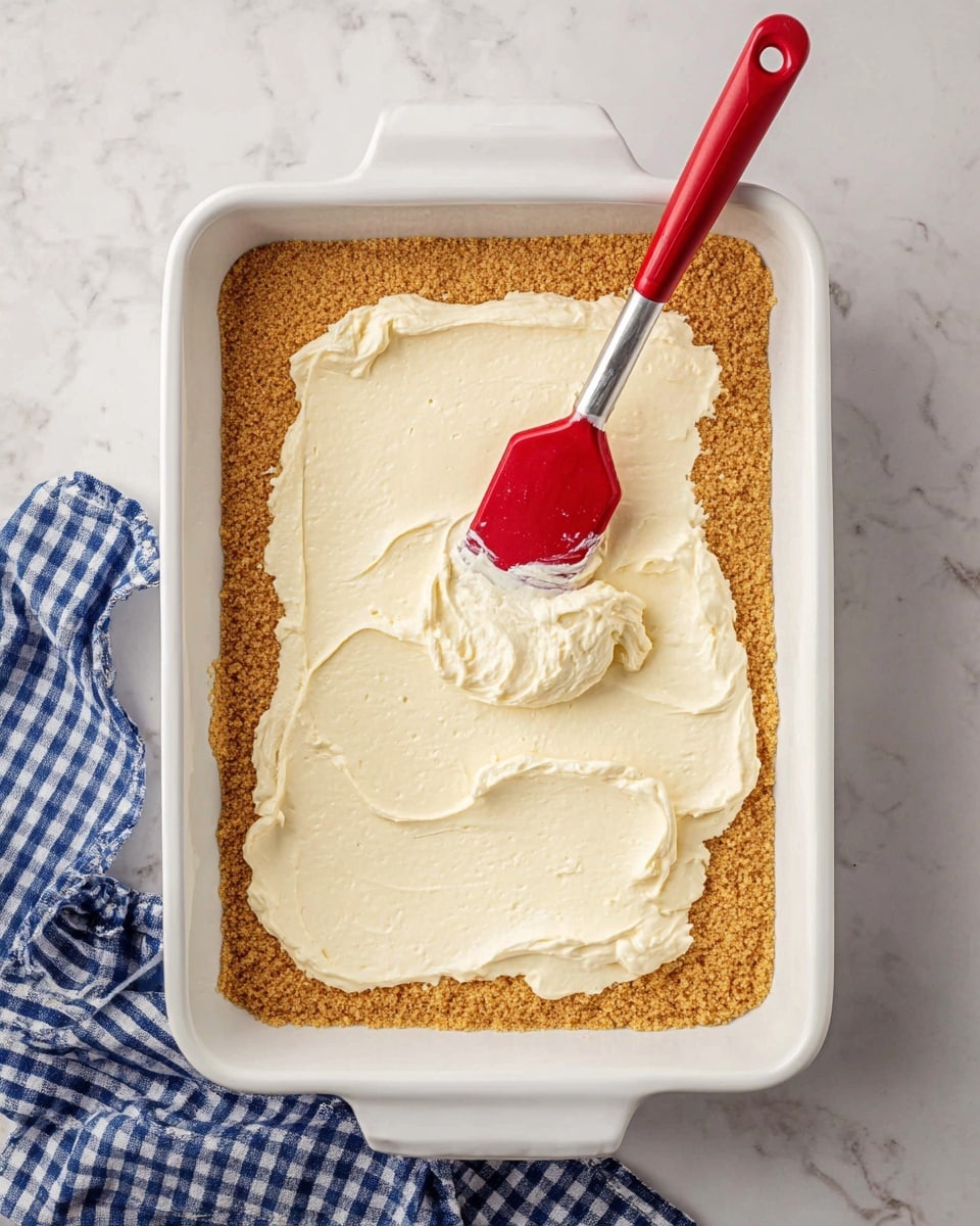 The image shows a white rectangular baking dish with a thick, even layer of golden brown crumb base covering the bottom. On top of this base, there is a second layer of creamy, smooth, off-white filling being spread with a red spatula, which has some of the filling on its surface. The baking dish is placed on a white marbled surface, and a blue and white checkered cloth is placed partially under the dish in the bottom right corner. Photo taken with an iphone --ar 4:5 --v 7