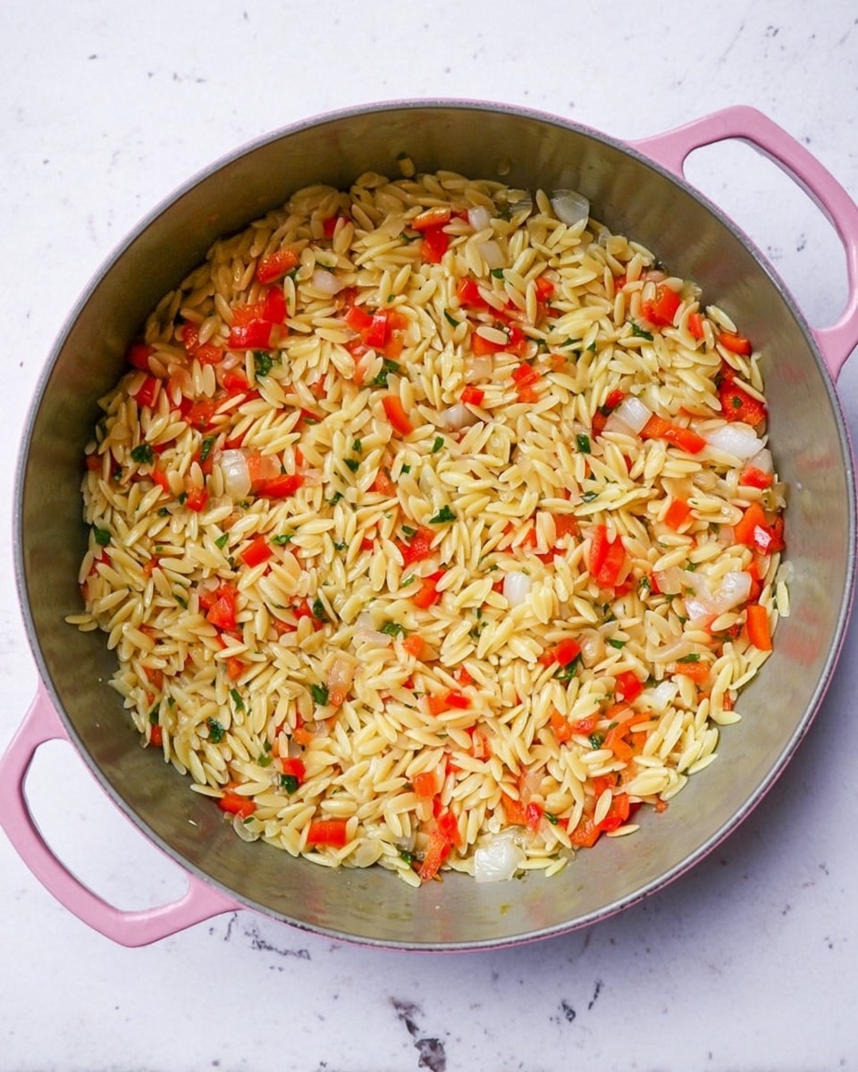 A gray pan with pink handles is filled with a single layer of cooked orzo pasta mixed evenly with small chopped pieces of red bell pepper and white onions. The orzo is light golden yellow, and the vegetables add bright red and translucent white colors throughout. The pan sits on a white marbled surface, with all ingredients well spread in the pan. photo taken with an iphone --ar 4:5 --v 7