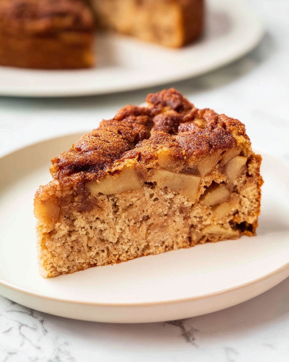 A single slice of apple cake sits on a plain white plate with a white marbled surface beneath. The cake has two layers visually: the bottom layer is a thick, light brown, moist cake base dotted with small pieces of cooked apple, while the top layer features a darker, cinnamon-spiced apple topping with a slightly crispy texture and uneven surface. The slice is triangular, and the cake's crumb looks soft and dense. The background shows a blurred white plate holding the remaining cake. Photo taken with an iphone --ar 4:5 --v 7