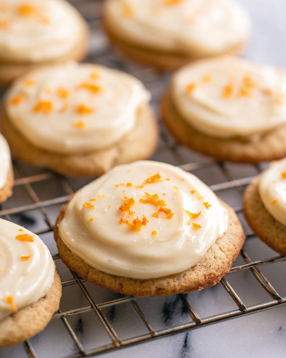 The image shows several round cookies with a beige-brown base layer visible at the bottom, topped with a creamy white frosting spread thickly but unevenly over each cookie. Small bright orange zest pieces are sprinkled lightly on top of the frosting, adding a pop of color. The cookies rest on a wire cooling rack over a surface with a white marbled texture. The focus is on the cookie in the center and the depth of field gently blurs the cookies in the background. Photo taken with an iphone --ar 4:5 --v 7