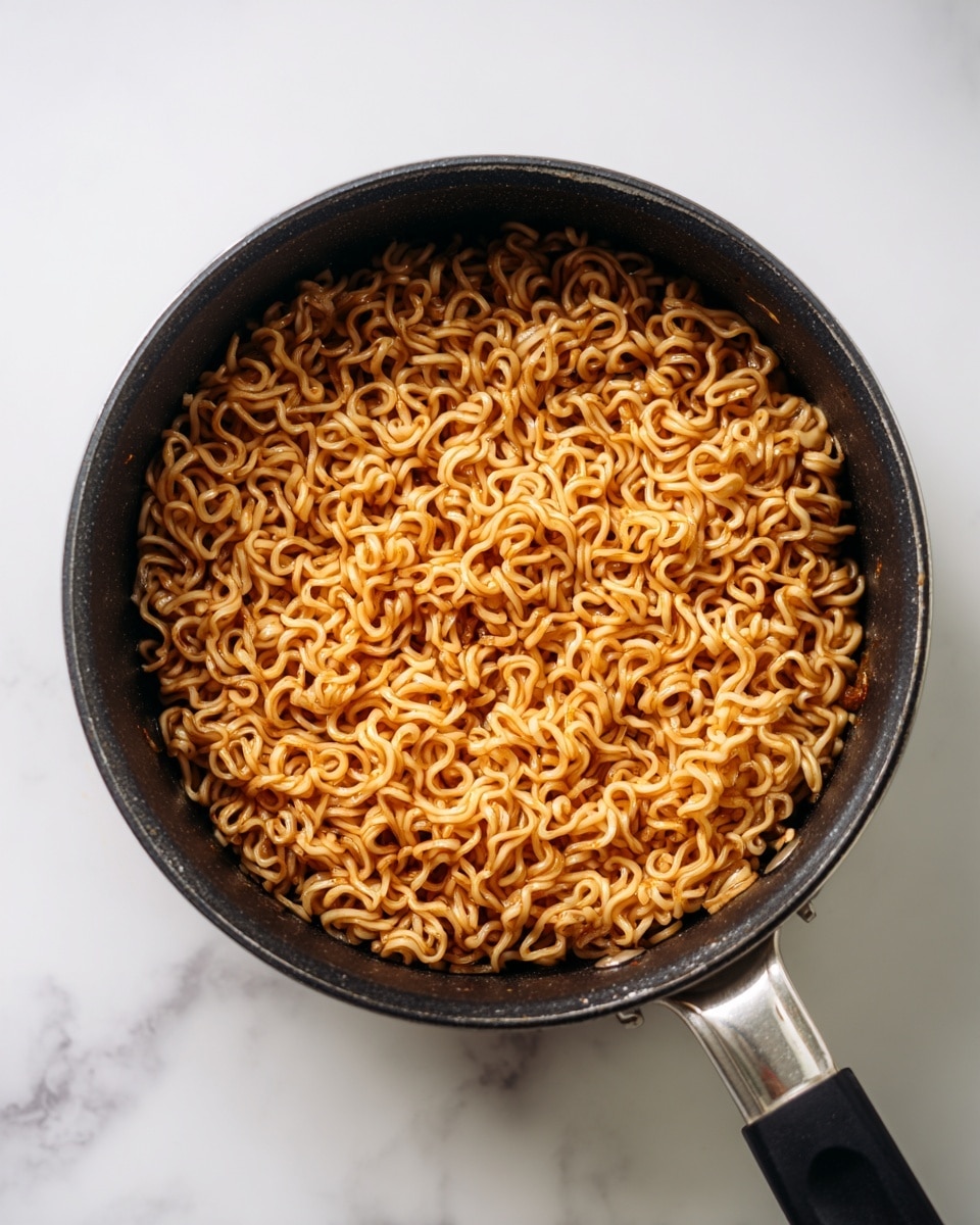 A black cooking pot filled with a single layer of cooked instant noodles, lightly coated with a thin, shiny brown sauce, showing tightly curled and slightly wavy strands evenly spread across the surface. The pot has a silver handle with a black grip, placed on a white marbled surface. The noodles look soft and moist with no additional toppings or garnish visible. photo taken with an iphone --ar 4:5 --v 7