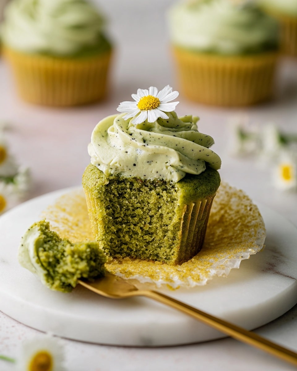 A small green cupcake with a soft, crumbly texture sits on a white marbled round surface. The cupcake has two layers: the bottom is a moist, dense green cake, and the top is thick, swirled light green frosting with tiny dark specks inside. On top of the frosting, there is a small white flower with a yellow center. The cupcake liner is open and spread under the cake, showing its yellowish inside. A gold spoon next to the cupcake holds a bitten piece of the cake with some frosting on it. There are some blurred cupcakes and small white flowers with yellow centers in the background. Photo taken with an iphone --ar 4:5 --v 7