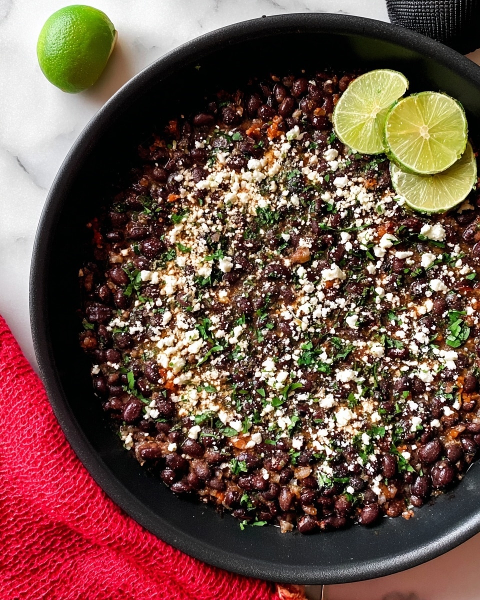 A black round pan holds a layer of cooked black beans mixed with small bits of orange and brown, topped with small white cheese crumbles and chopped green herbs scattered evenly across. Two lime slices rest near the top edge of the pan on the beans. The pan is placed on a white marbled surface next to a red textured cloth and a black object. Photo taken with an iphone --ar 4:5 --v 7