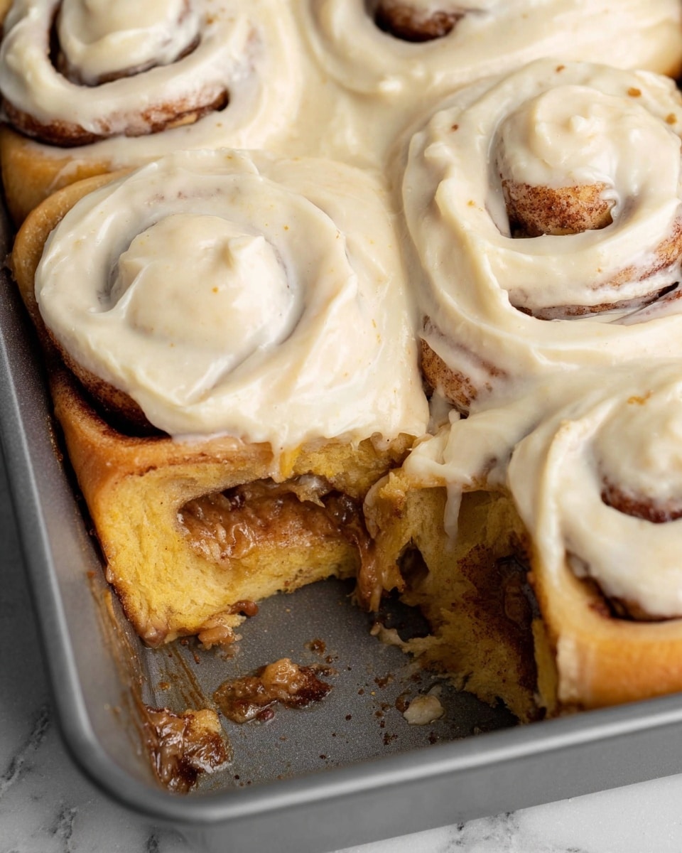 A close-up view of four cinnamon rolls in a metal baking pan on a white marbled surface, with the front right roll missing revealing its soft, fluffy, light golden inner layer and sticky syrupy bits underneath; each roll is topped with a thick, creamy, off-white frosting with tiny visible specks and a smooth yet swirled texture covering the tops in generous layers; the rolls show a slightly caramelized edge where they touch the pan and each other, with visible dark cinnamon swirls inside the dough; the pan has some syrup residue and the overall image focuses tightly on the textures and layers of the rolls photo taken with an iphone --ar 4:5 --v 7