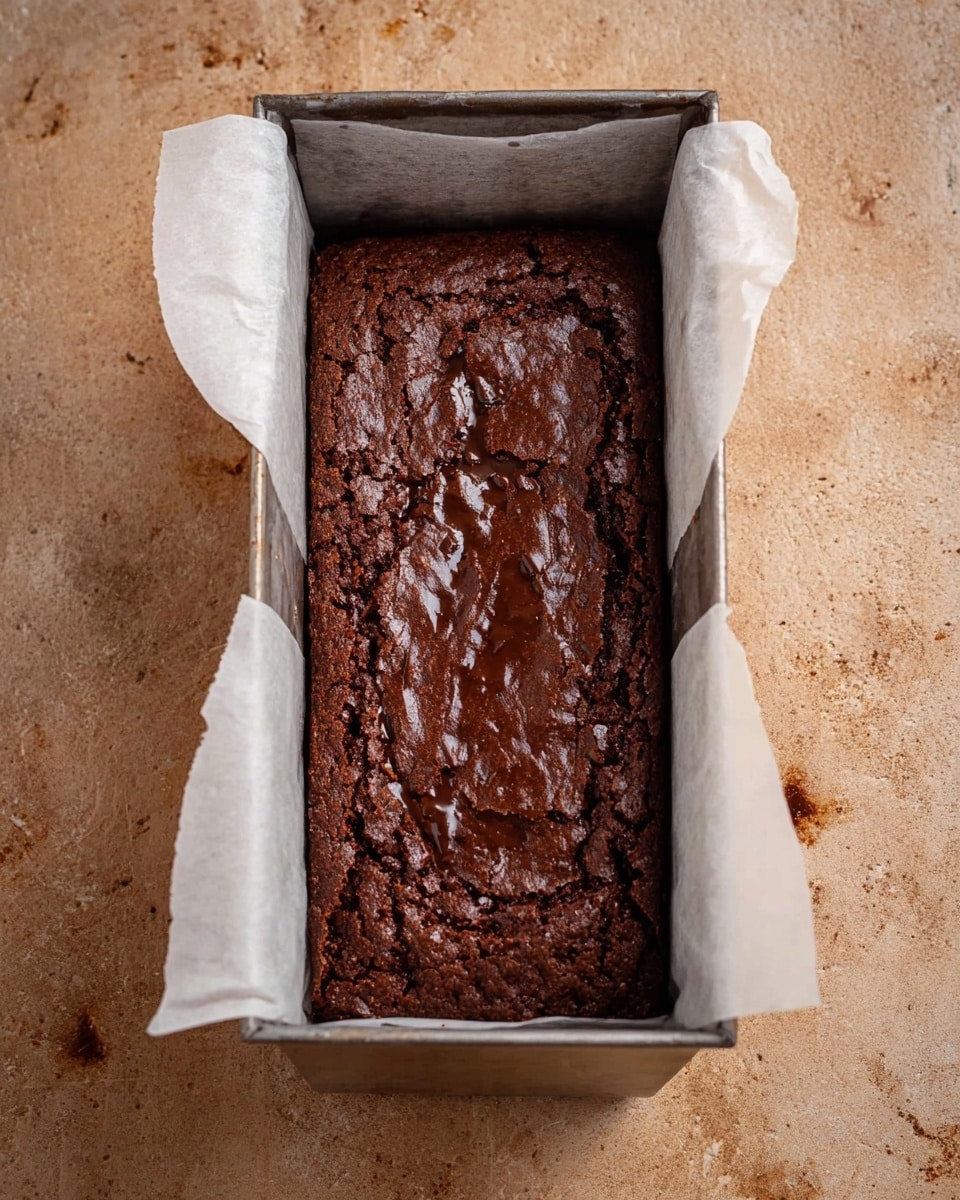 A rectangular dark brown chocolate brownie with a slightly shiny, crinkled top layer sits inside a metal loaf pan lined with white parchment paper on both long sides, the edges of the paper extending above the pan. The brownie has a textured surface with small cracks and dark spots indicating chunks of chocolate or nuts. The background is a light brown surface with small stains. photo taken with an iphone --ar 4:5 --v 7