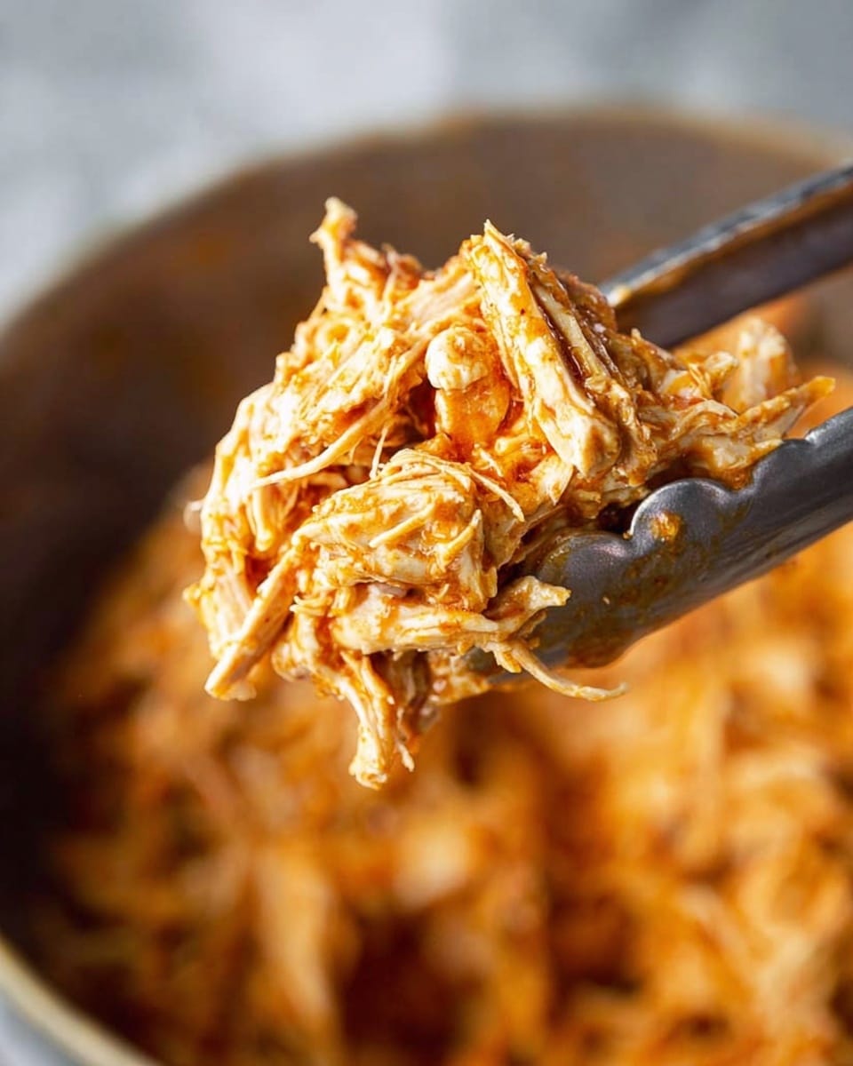 A close-up view of shredded chicken mixed with a bright orange sauce, held by metal tongs. The chicken pieces are fibrous and juicy, showing their tender texture. The background is blurred but suggests a large bowl filled with more of the same shredded chicken mixture. The lighting highlights the moist and slightly glossy surface of the chicken strands. The setting includes a white marbled texture surface. photo taken with an iphone --ar 4:5 --v 7