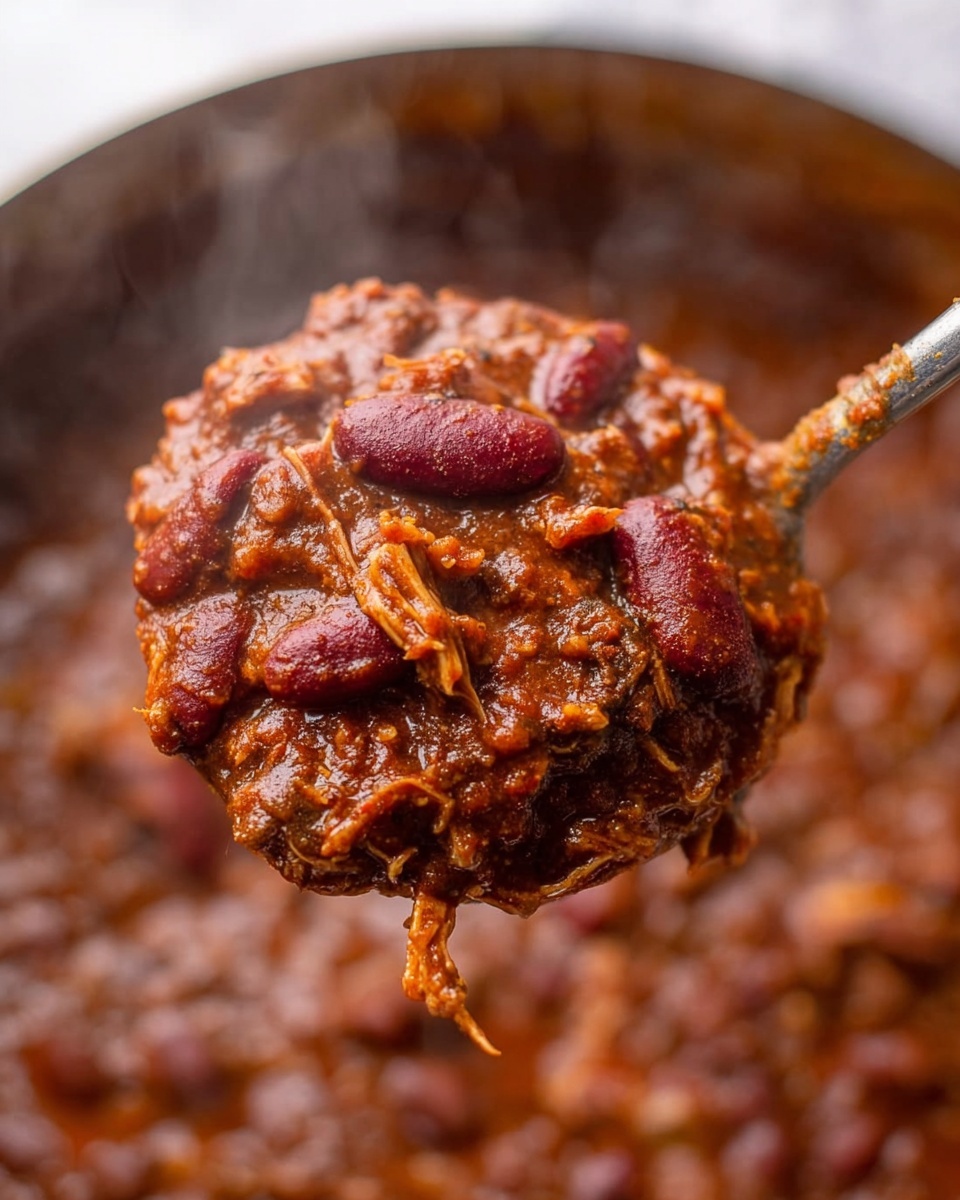 A close-up view of a large spoon holding thick chili with dark red kidney beans and shredded tender meat in a rich, dark brown sauce. The chili is textured with visible chunks of tomatoes and spices, creating a hearty and steamy appearance. The background shows more chili in the pot with a soft focus, and the whole scene rests on a white marbled texture. photo taken with an iphone --ar 4:5 --v 7